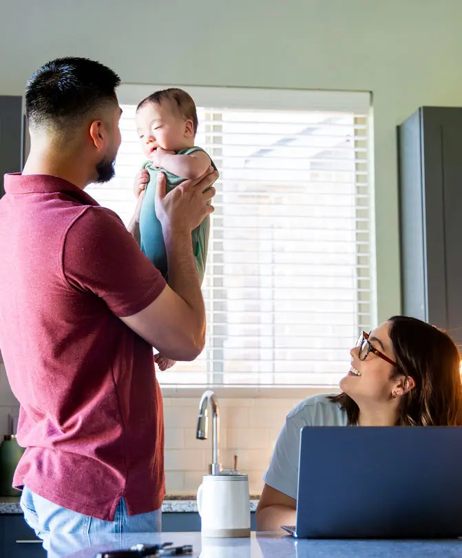 man-holding-a-baby-in-a-kitchen-while-a-woman-works-on-a-laptop