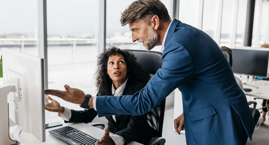 man-leaning-over-a-seated-woman’s-desk-in-an-office